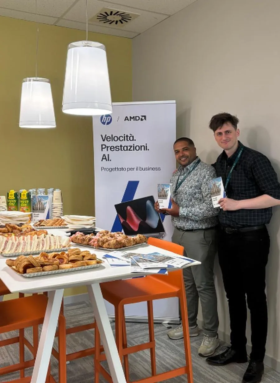 Two colleagues at the Italian event, each holding a copy of the Italian edition, standing beside an HP and AMD roller banner reading Velocità. Prestazioni. AI. Further copies are fanned out on the table in front of them