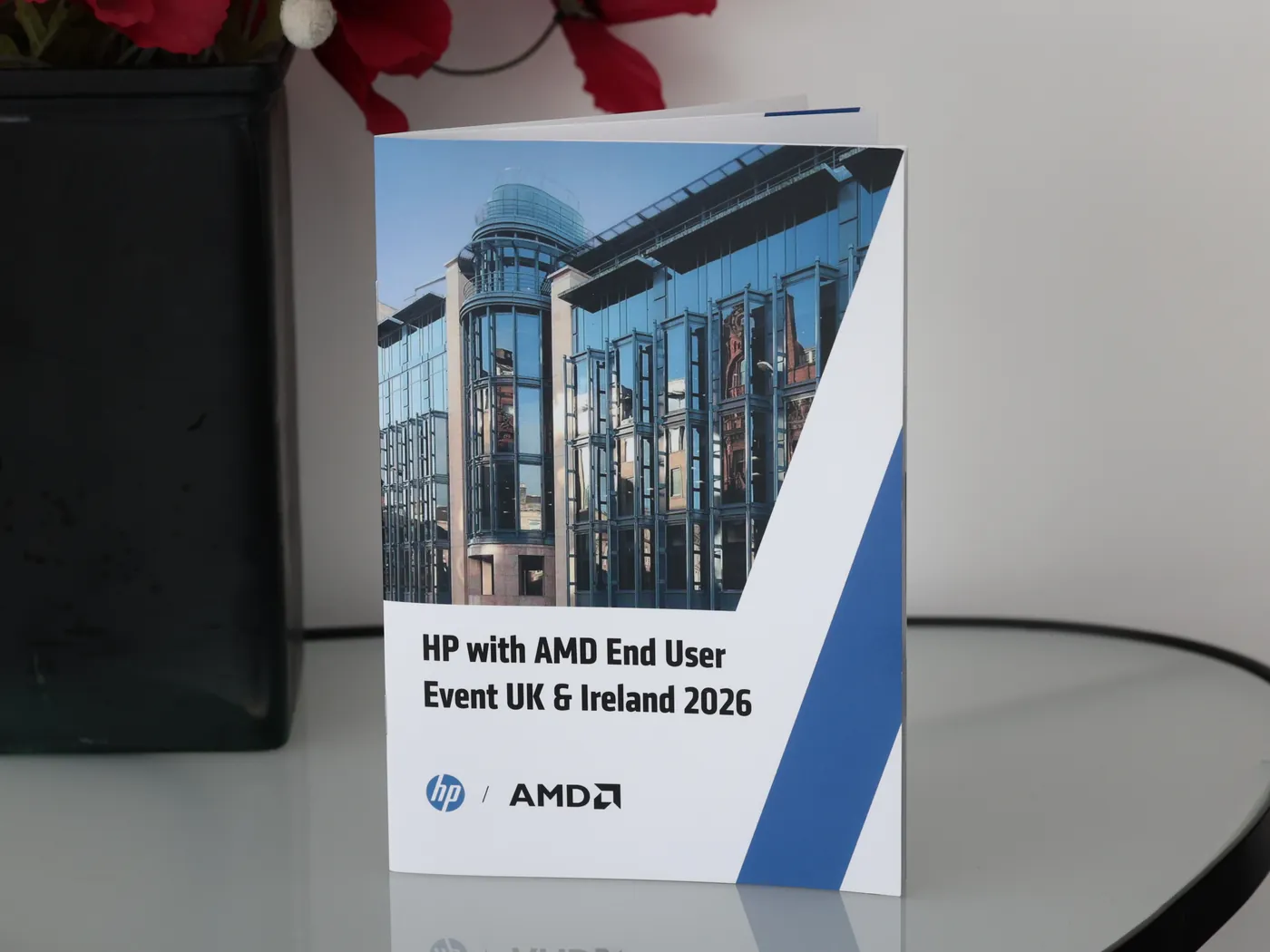 The printed HP with AMD End User Event booklet standing on a glass table beside a vase of red poppies