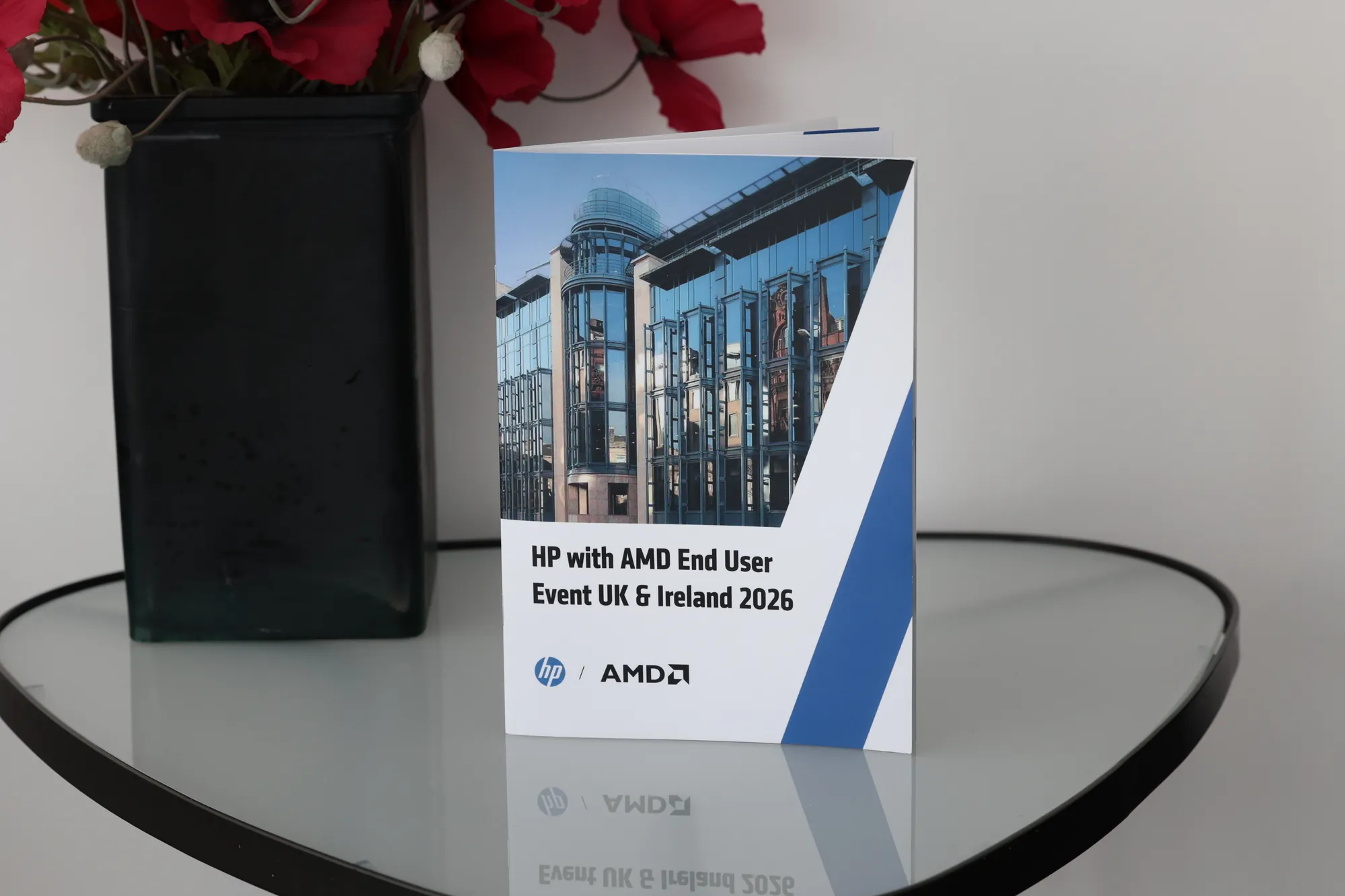 The printed first edition standing on a glass table beside a vase of red poppies: the cover reads HP with AMD End User Event UK and Ireland 2026, over a photograph of a glass office façade cut by a blue diagonal stripe