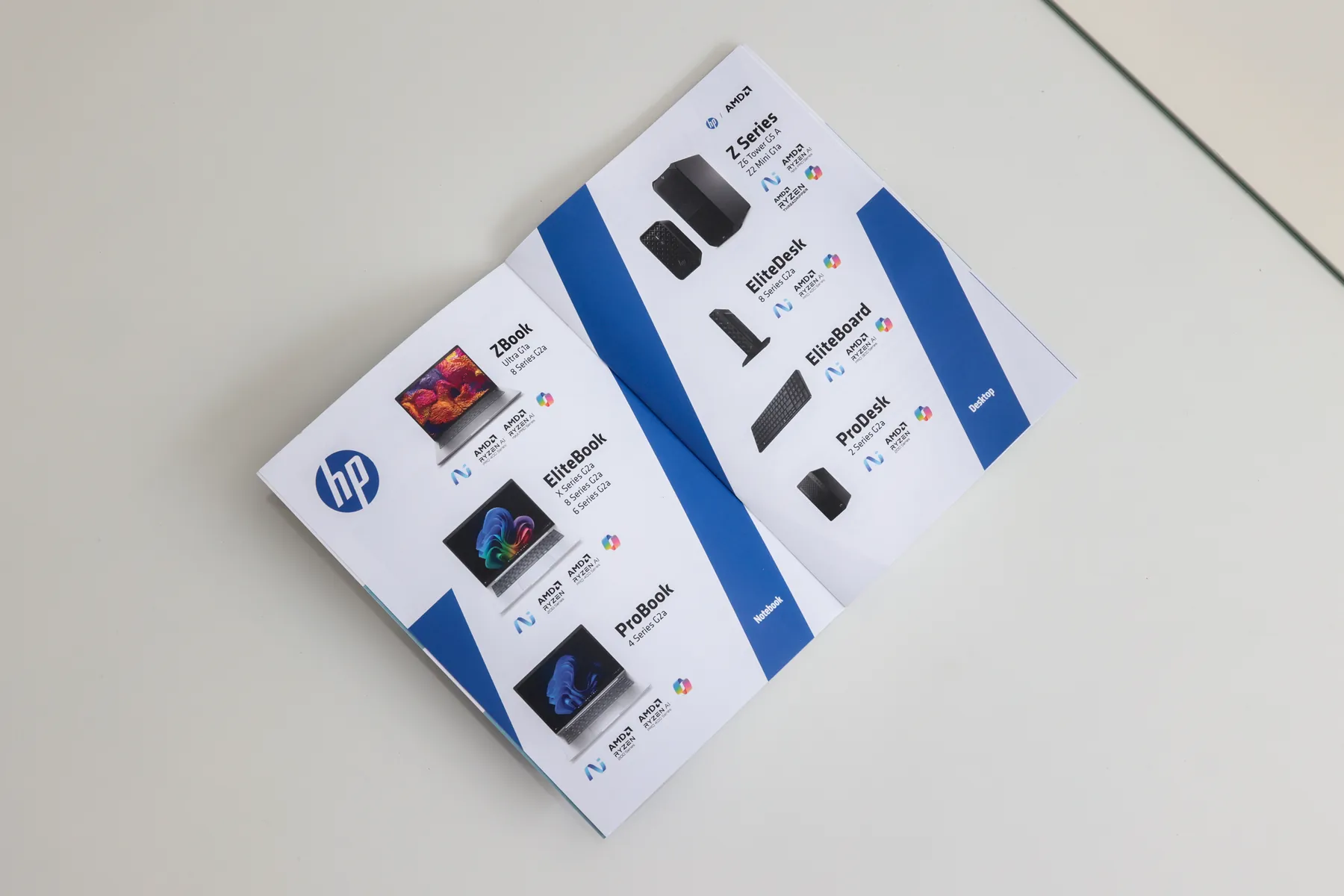 The booklet lying open, photographed at an angle, showing the notebook range on the left page and the desktop range on the right, each product tagged with its AMD Ryzen and Copilot badges