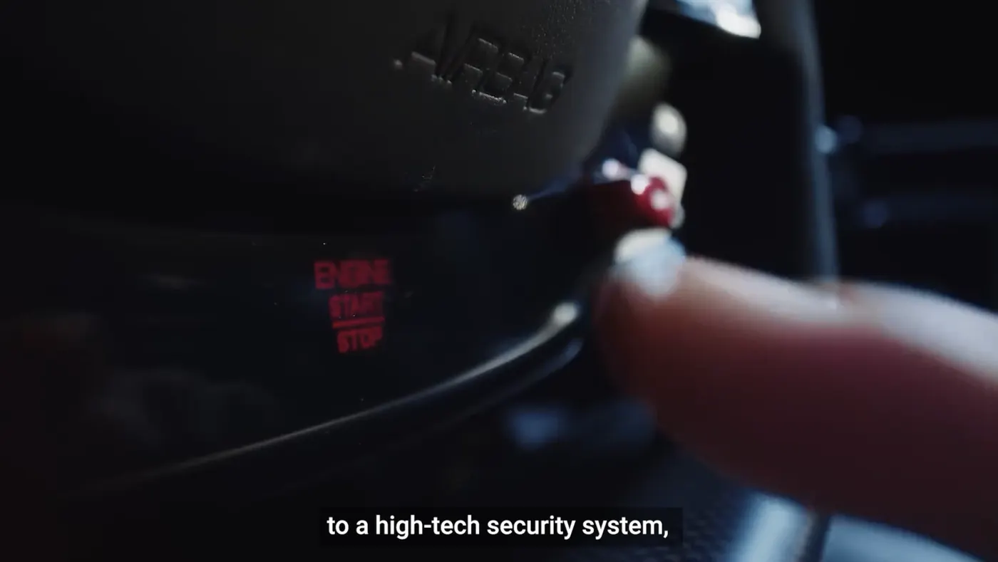 Close on a finger pressing an engine start-stop button glowing red beside an airbag cover, subtitled to a high-tech security system